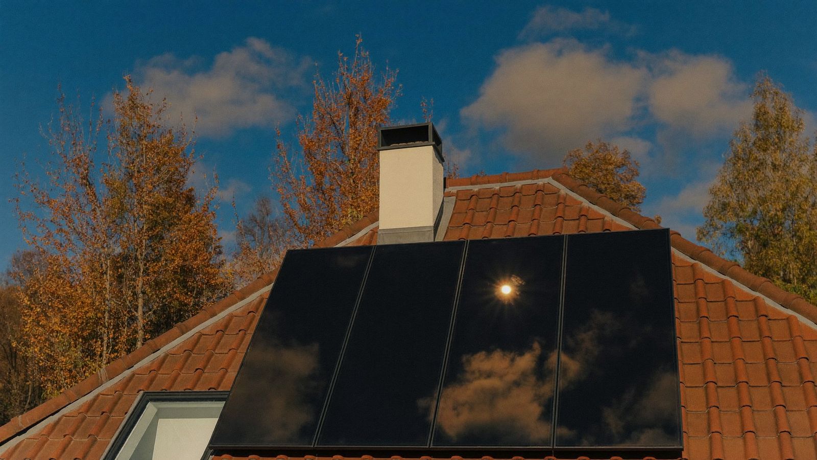 Fyra stående solpaneler med blankt svart glas som speglar sol och moln på ett rött tegeltak, med vit skorsten och gula höstträd mot blå himmel